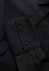 Poignet de chemise noire avec deux boutons noirs et coutures visibles sur tissu lisse.