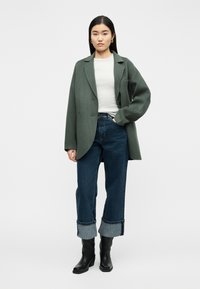 Γυναίκα στέκεται φορώντας σκούρο πράσινο φαρδύ σακάκι, λευκό τοπ, σκουρό μπλε τζιν με γυρισμένα μπατζάκια, μαύρες μπότες, με το ένα χέρι στην τσέπη, μπροστά σε απλό φόντο.