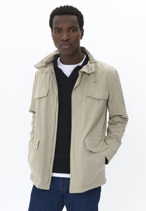 Giacca impermeabile beige con cappuccio, dotata di due tasche sul petto, tasche laterali e chiusura con zip. Indossata sopra un maglione nero e una camicia bianca.