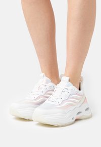 Sneaker bianca con accenti arcobaleno, tomaia in rete e materiali sintetici, suola spessa, motivi testurizzati e chiusura con lacci. Nessun marchio visibile.