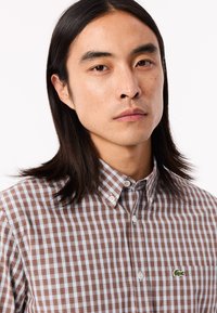 Camisa de cuadro marrón y azul claro con botones y cuello. Presenta un pequeño logo de cocodrilo verde en el pecho. Tela de algodón texturizada.