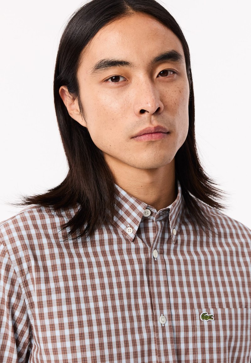 Chemise à carreaux marron et bleu clair avec col. Présente un petit logo crocodile vert sur la poitrine. Tissu en coton texturé.