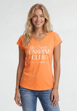 Young woman with long blonde hair wearing an orange t-shirt with white text and blue jeans, smiling against a white background.