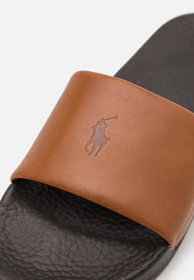 Sandal de tiras em couro castanho com sola preta texturizada. Apresenta um logótipo em relevo na tira. O design enfatiza o conforto e o uso casual.