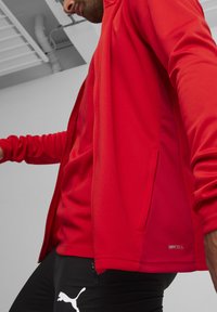 Rote Sportjacke mit glatter Textur und Reißverschluss, ausgestattet mit Netzlüftern und geprägtem Branding, kombiniert mit schwarzen Hosen.