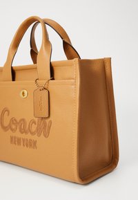 Karamell bőr Coach kézitáska dupla fogantyúkkal, külső zsebbel, arany színű zárókapcsolóval és beleégett "Coach New York" márkajelzéssel.