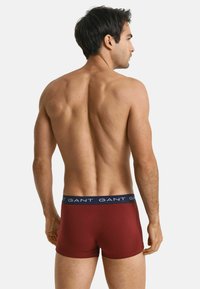 Herren-Boxershorts aus tiefrotem Stoff mit einem dunkelblauen Bund, der mit wiederholt aufgedrucktem Logo-Text versehen ist; enganliegende Passform, mittellange Beine und glatte Textur.