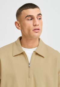 Beige ritssluiting pullover met een grote kraag, gemaakt van zacht materiaal. Heeft een zilveren rits en een witte onderhemd zichtbaar eronder.