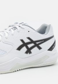 Zapatilla deportiva ASICS blanca y gris con logo negro, cordones blancos, suela acolchada y superficie superior texturizada, mostrada desde un ángulo lateral.