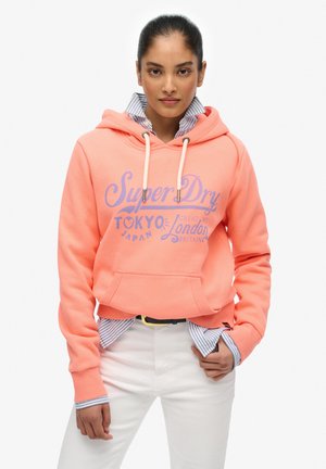 Sudadera corta color coral con un bolsillo frontal, cordones blancos y texto gráfico púrpura; superpuesta sobre una camisa a rayas azul y blanca.