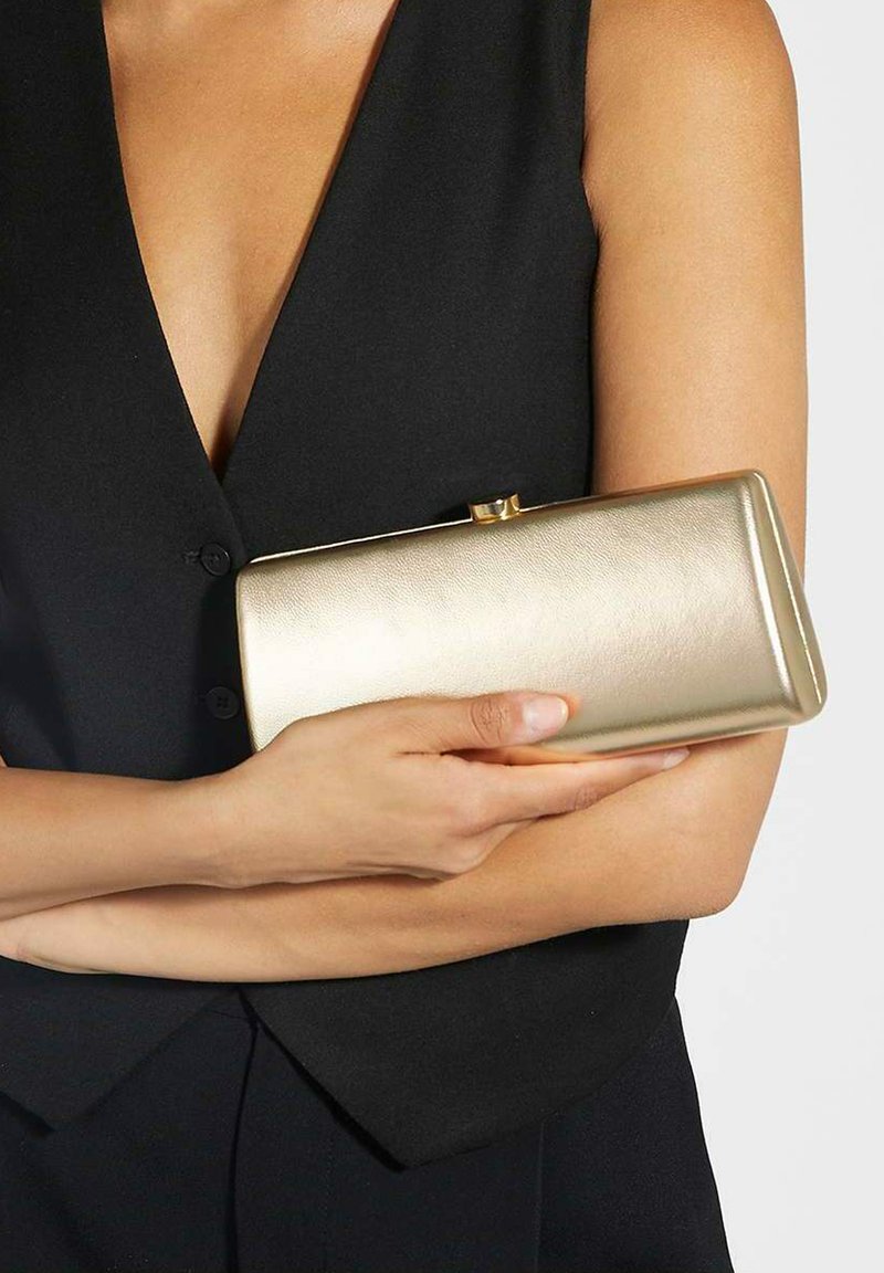 Gold metallic clutch with a smooth texture, rectangular shape, and a clasp closure, held against a black garment.