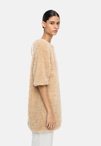 Beżowa, sztuczna futrzana bluzka w oversize'owym fasonie z krótkimi rękawami, teksturowanym wykończeniem, zaokrąglonym dołem i otwartym tyłem. Stylizowana z jasnymi spodniami.