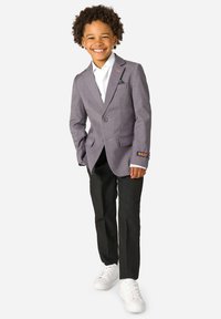 Uśmiechnięte dziecko z kręconymi włosami, ubrany w szary żakiet, białą koszulę, czarne spodnie i białe trampki, stojące na tle jednolitego tła.