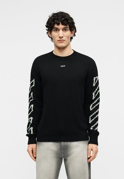 Νεαρός άνδρας που φοράει μαύρο πουλόβερ με λευκό κείμενο "OFF" στο στήθος και λευκά ζιγκ-ζαγκ μοτίβα στα μανίκια, συνδυασμένο με ανοιχτό γκρι τζιν.