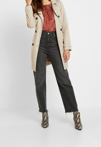 Trench coat beige con bottoni neri, abbinato a un top color ruggine e jeans a vita alta grigio scuro. Stivali alla caviglia con stampa pitone.
