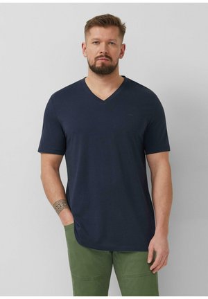 Homme aux cheveux courts et barbe, portant un T-shirt bleu marine à col en V et un pantalon vert, debout devant un fond gris clair uni.