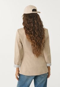 Blazer beige su misura con maniche lunghe, polsini a costine e due tasche laterali, abbinato a un cappellino da baseball beige chiaro. Jeans in denim casual.