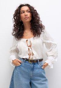 Femme aux cheveux bouclés portant une blouse blanche à manches longues avec des liens et un jean bleu taille haute, les mains dans les poches, regardant sur le côté.