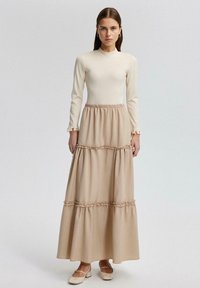 Lange beige jurk met een aansluitende, lange mouw en een tiered frilzoom, gemaakt van soepele stof, met subtiele gerimpelde manchetten en een elastische taille.
