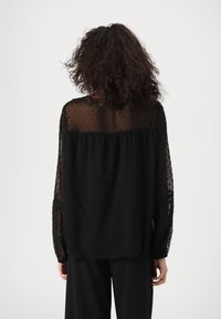Blusa nera con un tessuto trasparente a pois, maniche lunghe e vestibilità ampia. Il retro presenta un design arricciato per una maggiore texture.