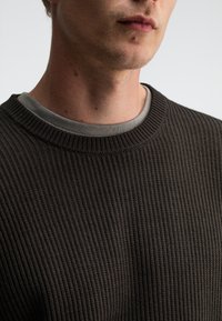 Dunkel gestreifter Strickpullover mit Rundhalsausschnitt, über einem hellgrauen Hemd getragen. Die Textur ist ausgeprägt mit einem strukturierten, gewebten Erscheinungsbild.