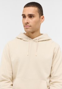 Beżowa bluza z kapturem wykonana z miękkiego materiału, z przednią kieszenią, kapturem ze sznurkiem i subtelnym logo na piersi.