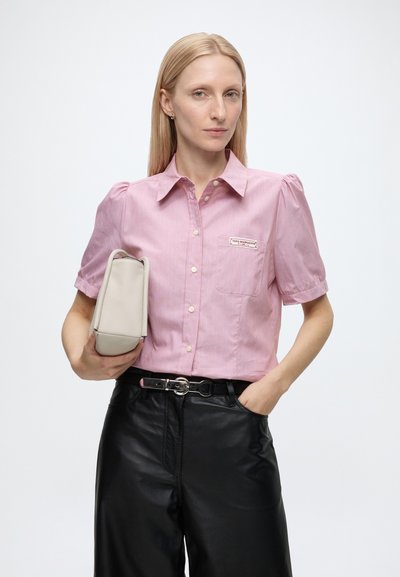 Femme aux cheveux blonds et raides portant une chemise rose à manches courtes avec boutons et un pantalon noir, tenant une pochette beige sous le bras.