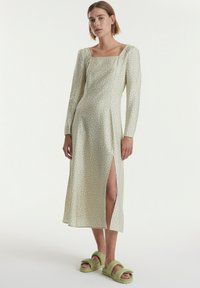 Langes, blassgrünes Kleid mit quadratischem Ausschnitt und weißen Punkten; verfügt über lange Ärmel und einen Schlitzausschnitt. Getragen mit hellgrünen Slides.