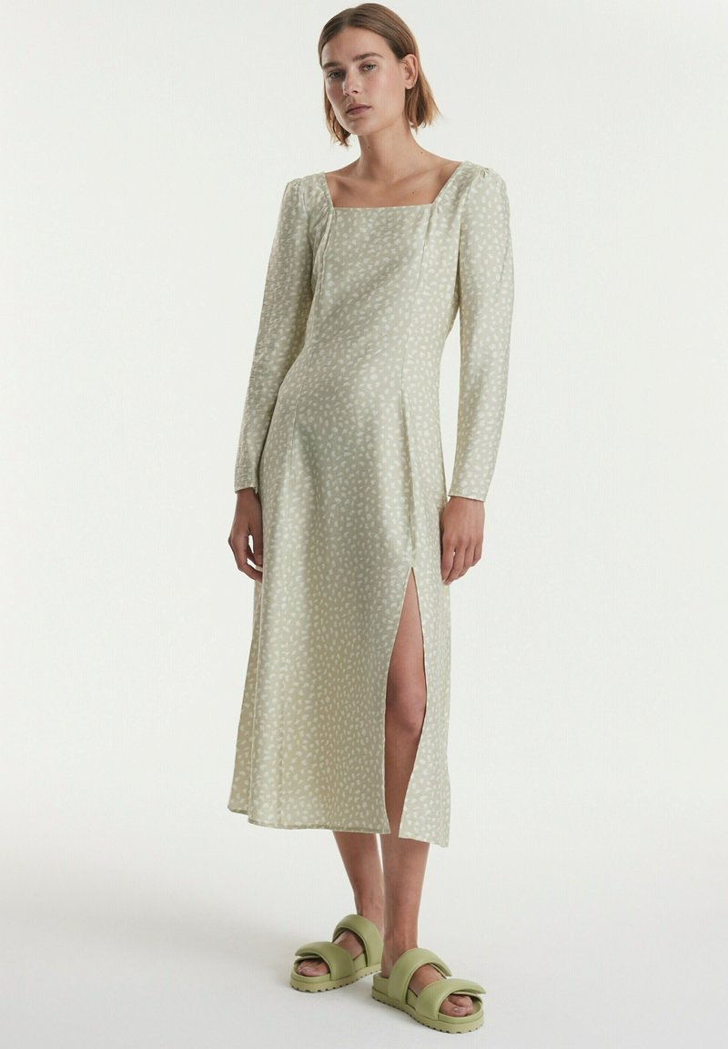 Langes, blassgrünes Kleid mit quadratischem Ausschnitt und weißen Punkten; verfügt über lange Ärmel und einen Schlitzausschnitt. Getragen mit hellgrünen Slides.