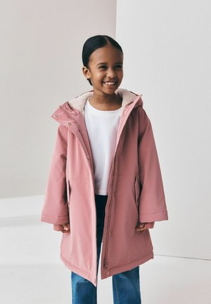 Ein junges Mädchen steht lächelnd da und trägt einen langen pinken Mantel über einem weißen Hemd und blauen Jeans vor einfachem, hellem Hintergrund.