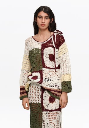 Mujer joven vistiendo un suéter y una falda de ganchillo patchwork en tonos crema, verde oliva, granate y amarillo, de pie frente a un fondo liso.