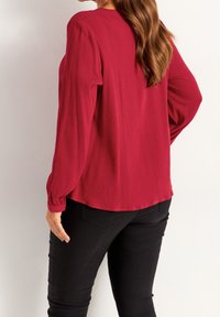 Top lungo rosso con finitura testurizzata; vestibilità comoda, scollo rotondo e polsini arricciati. Abbinato a pantaloni neri aderenti.