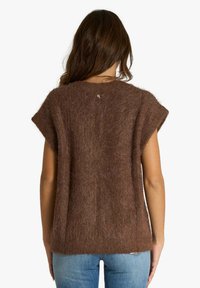 Maglione marrone peloso con maniche corte, trama spessa, vestibilità ampia e piccolo logo sul retro del collo. Indossato sopra jeans azzurri.