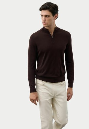 Uomo che indossa un maglione marrone scuro con zip a un quarto e pantaloni color panna, che guarda a sinistra su uno sfondo chiaro e semplice.