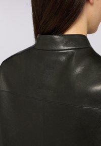 Giacca di pelle nera con una texture liscia e un colletto strutturato, caratterizzata da un logo discreto e dettagli di cucitura pulita sul retro.