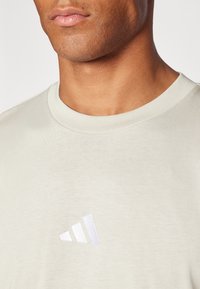 Světle šedé bavlněné tričko s kulatým výstřihem a malým bílým vyšitým logem Adidas na levé straně. Měkká textura a jednoduchý design.