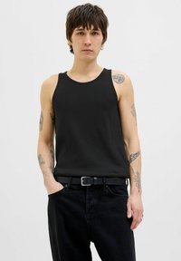 Débardeur noir à côtes avec des bretelles larges, porté avec un jean noir taille haute et une ceinture noire. Présente des tatouages visibles sur les bras.