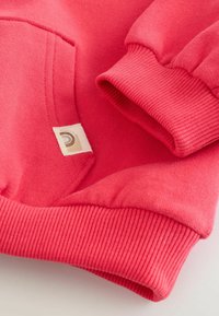 Sweatshirt rose corail avec poignets et ourlet côtelés, comportant une petite étiquette en tissu sur la poche. Texture douce et design décontracté.