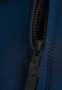 Közelkép egy fekete műanyag cipzárról, textil húzófüllel, amely egy sötétkék varrott ruhadarabon vagy táskán található.