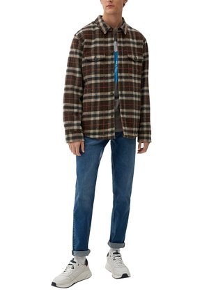 Braun-grün-creamfarbener karierter Flanellhemd mit zwei Brusttaschen, über einem grauen Grafik-T-Shirt getragen, kombiniert mit blauen Jeans und weißen Sneakers.