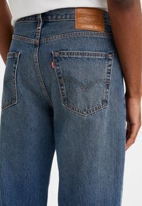Vue de dos d'une personne portant un jean en denim bleu Levi's avec un patch de marque visible et la célèbre étiquette rouge Levi's sur la poche arrière droite.