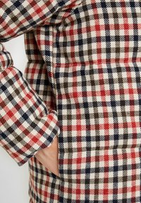 Tissu en mélange de laine à motif écossais rouge, bleu marine et marron ; comprend une poche et une surface texturée ; gros plan sur la manche et le côté.