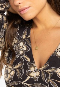 Collar de oro presentado en una modelo, con una blusa de patrón floral en tonos negro, crema y dorado, que muestra un diseño de escote en V.