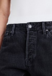 Jeans de denim negro con una textura ligeramente desvaída, con un cierre de botón plateado y costuras resistentes. No hay bolsillos visibles en el primer plano.