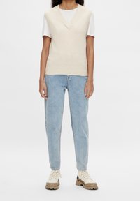 Personne portant un pull sans manches en tricot crème par-dessus un t-shirt blanc, un jean bleu clair et des bottes beiges à semelle épaisse, debout sur un fond blanc.