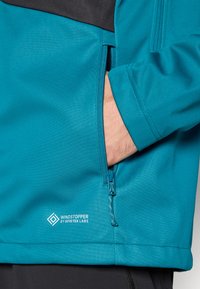Turkusowa kurtka Windstopper z czarną górną częścią. Posiada boczną kieszeń na zamek i teksturowany materiał. Logo widoczne na dole.
