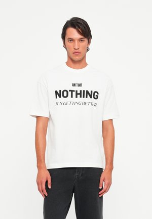 Young man with dark hair wearing a white t-shirt with black text reading "AIN'T GOT NOTHING IT'S GETTING BETTER" and black pants.
