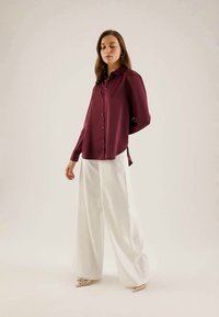 Burgundy színű, hosszú ujjú ing gombos záródással, széles fehér nadrággal párosítva. A modell egy semleges háttér előtt áll.