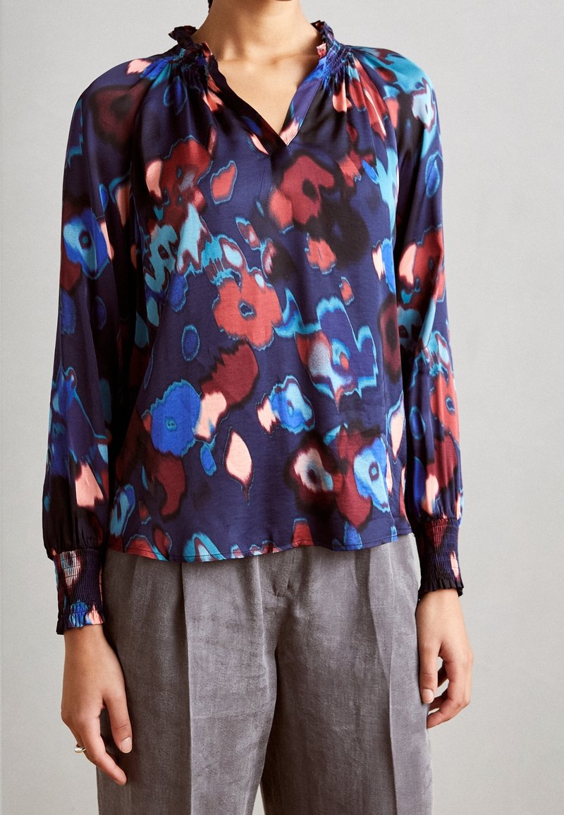 Femme portant un chemisier à manches longues avec un motif floral abstrait multicolore et un pantalon gris, debout devant un fond uni.