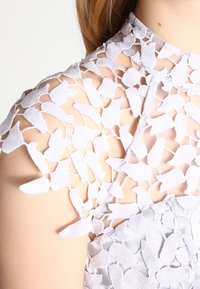 Hennon vaaleanlila mekko, jossa on yksityiskohtainen kukkakoristeinen pitsi olkapäillä, korkea kaula-aukko ja teksturoitu kangas.